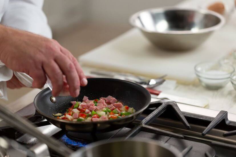 Chef adding ham to vegetables in a frying pan 